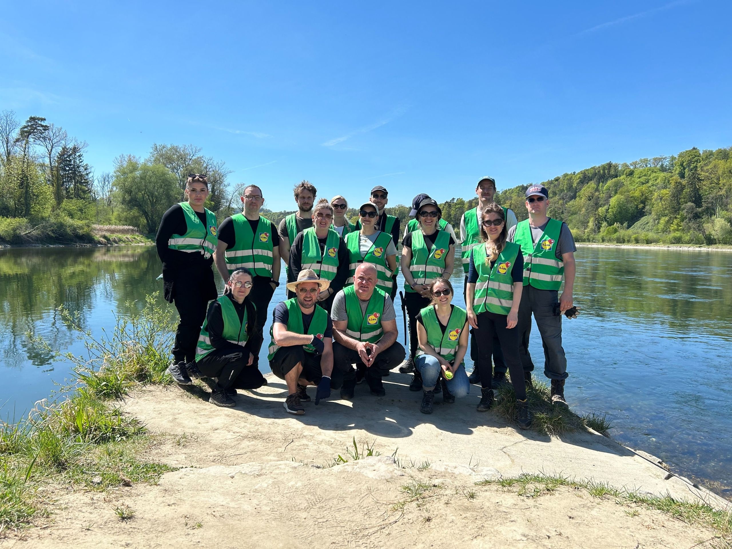 Onze collaborateurs de Lidl Suisse en gilets de sécurité verts posant en deux rangées sur un banc de sable au bord d'une rivière naturelle – photo d'équipe après l'engagement commun pour valoriser des habitats précieux pour le sonneur à ventre jaune et d'autres espèces protégées en Suisse.