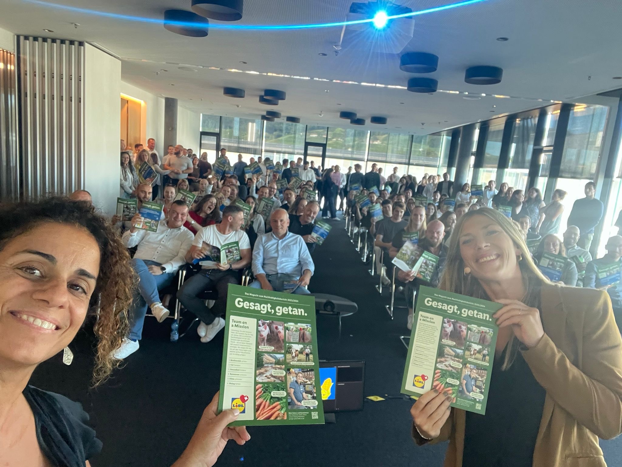Grazia Grassi und Milena Herrmann vor Mitarbeiter:innen, halten je eine Ausgabe des Magazins Gesagt, getan in die Kamera.  