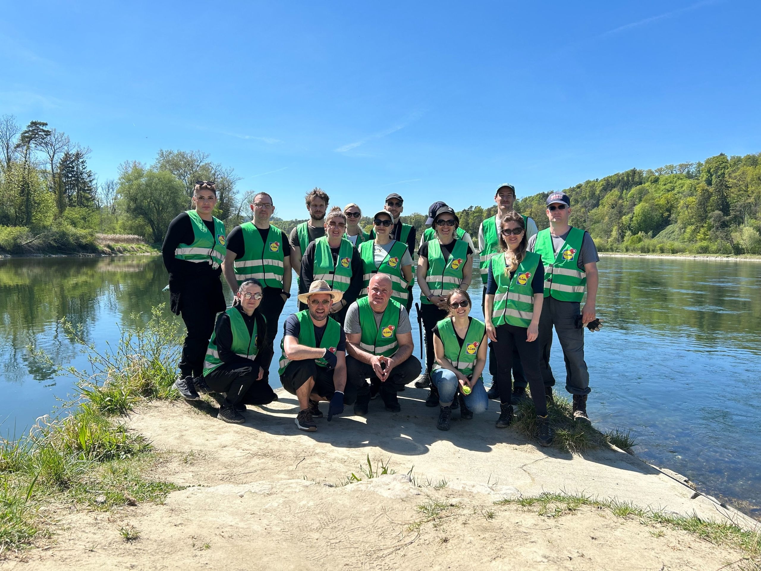 Undici dipendenti di Lidl Svizzera in gilet ad alta visibilità verdi posano in due file su un banco di sabbia lungo un fiume naturale – foto di gruppo dopo l'impegno comune per valorizzare habitat preziosi per l'ululone dal ventre giallo e altre specie protette in Svizzera.
