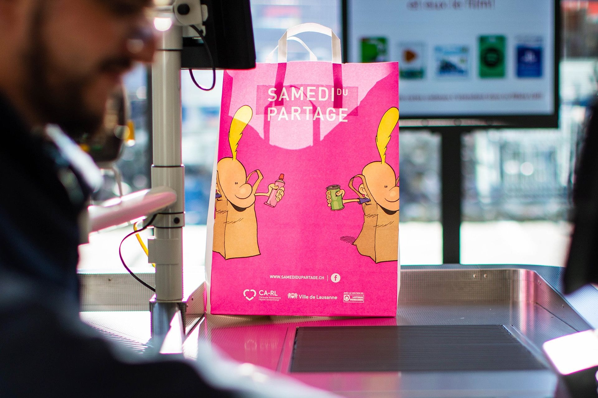 Le sac «Samedi du partage» se trouve au bout du tapis roulant de la caisse tandis qu'un employé continue de scanner les articles