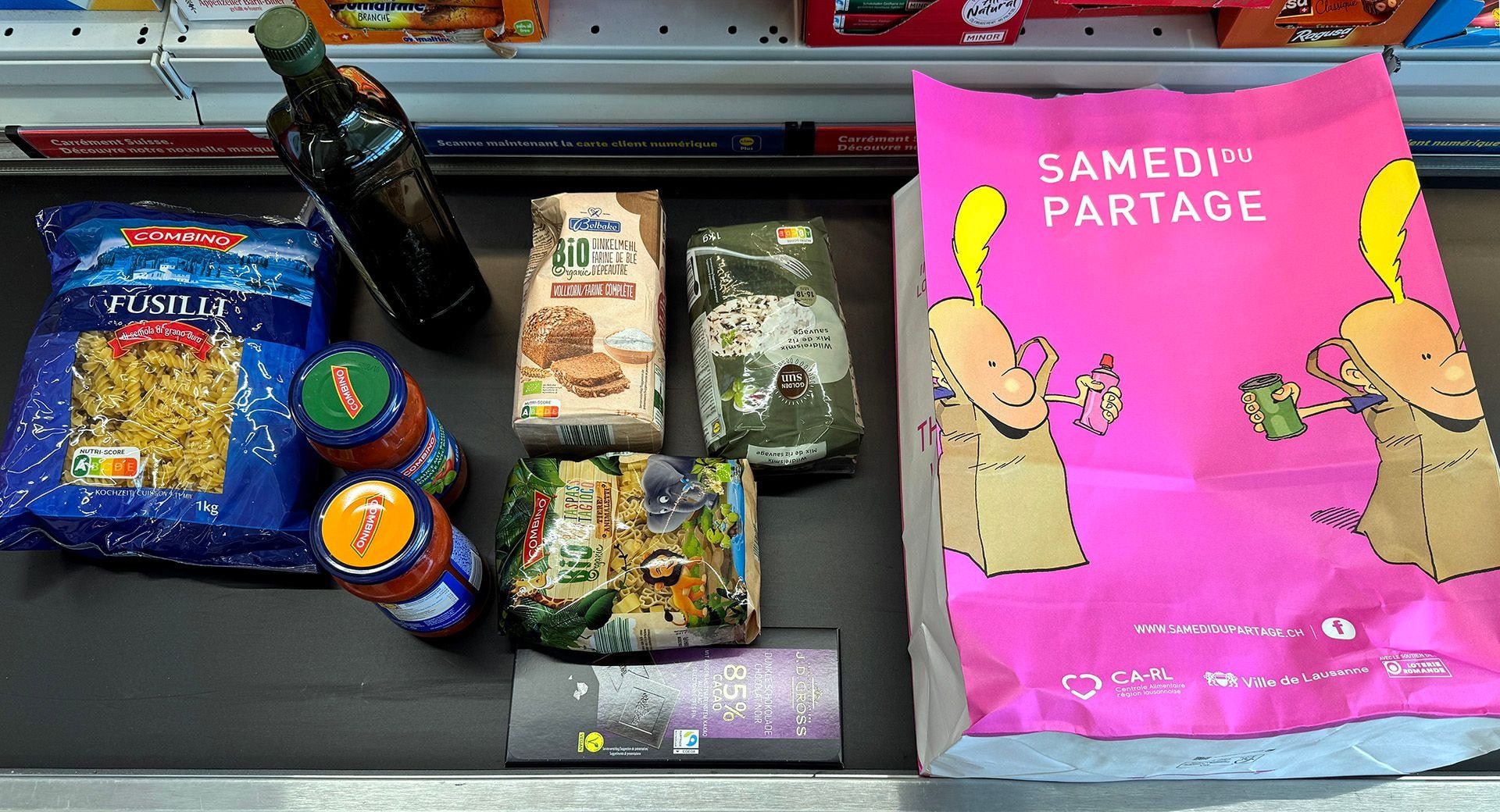 Auf einem Förderband liegen eingekaufte lidl Produkte, wie Pasta, Pesto, Schokolade und eine samedi du partage Einkaufstasche in pink