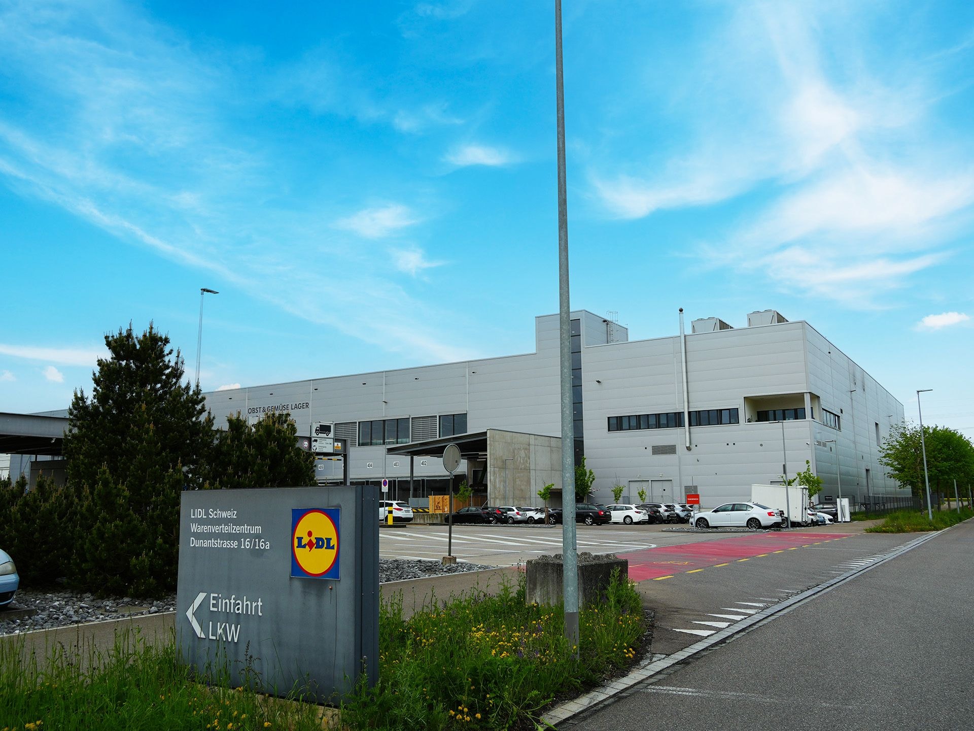 Blick auf den Parkplatz Lidl Schweiz warenverteilungszentrum, das Gebäude ist gross und eckig und hat eine aluminiumartige aussenerscheinung