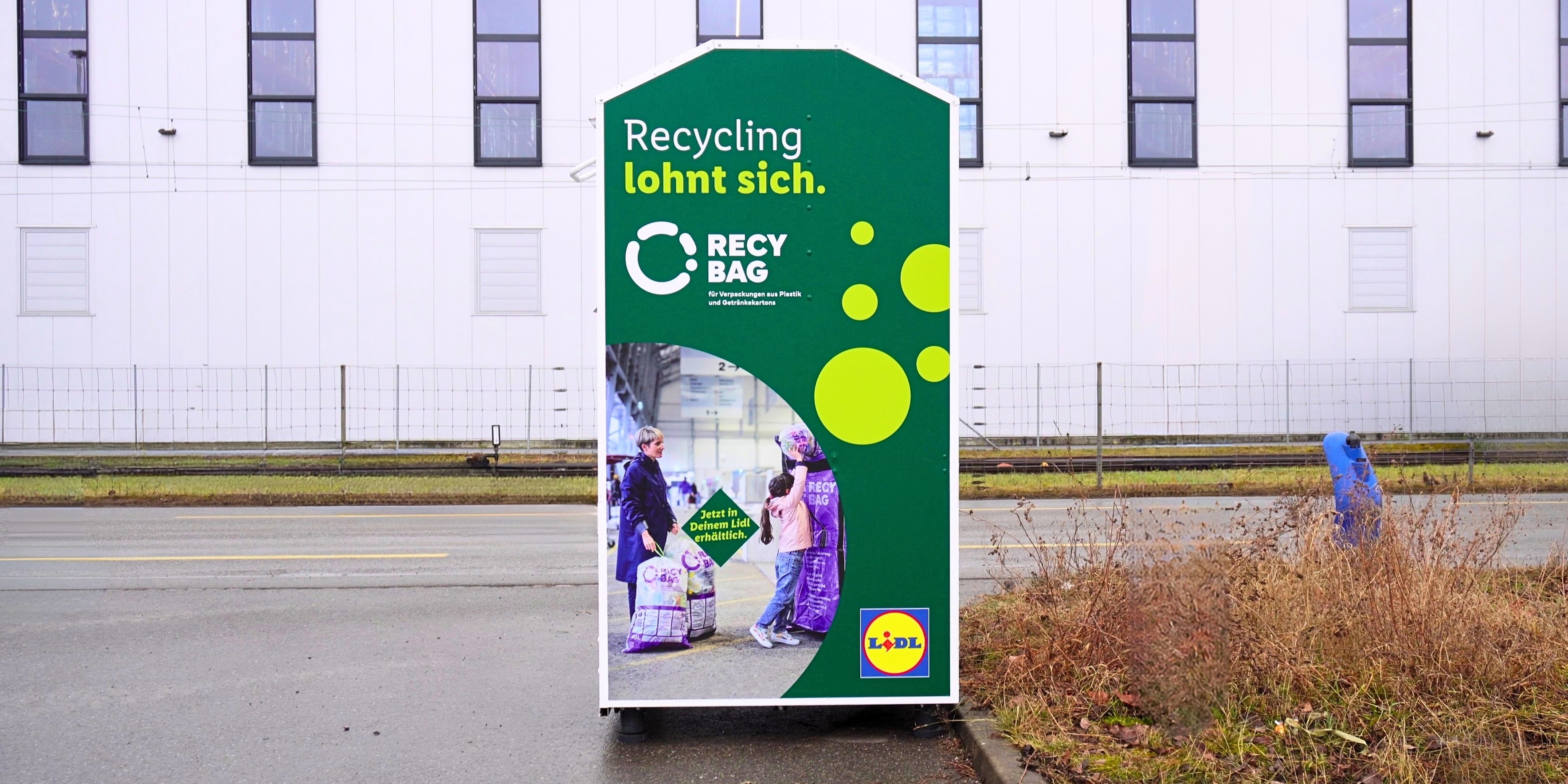 Weisser Lidl-Recycling-Container für „RecyBags“ auf einem Parkplatz vor einem Gewerbegebäude.