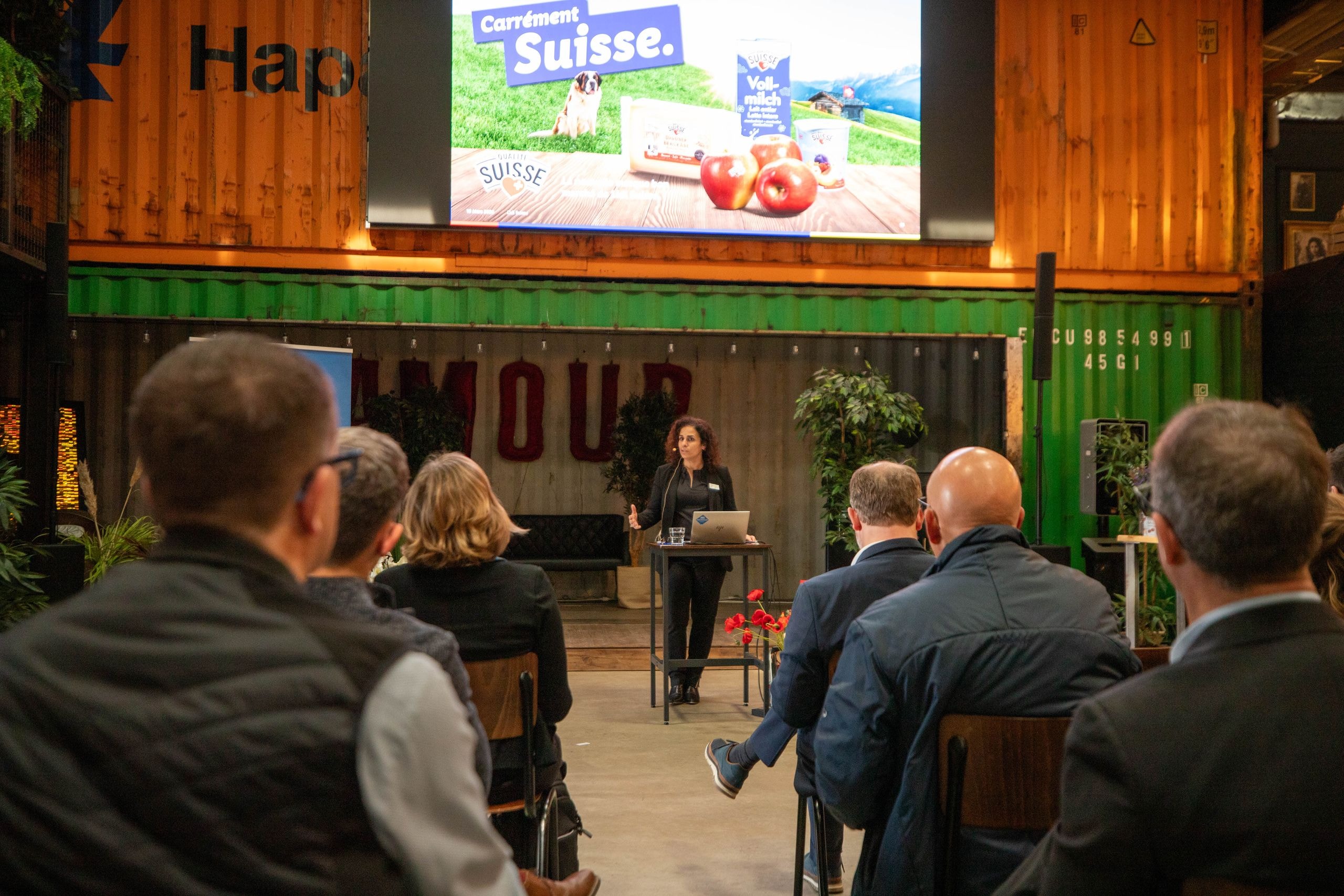 Eine Referentin präsentiert auf der Bühne vor Teilnehmenden des Stakeholder-Dialogs, im Hintergrund eine Projektion mit Schweizer Produkten und dem Slogan Carrément Suisse.