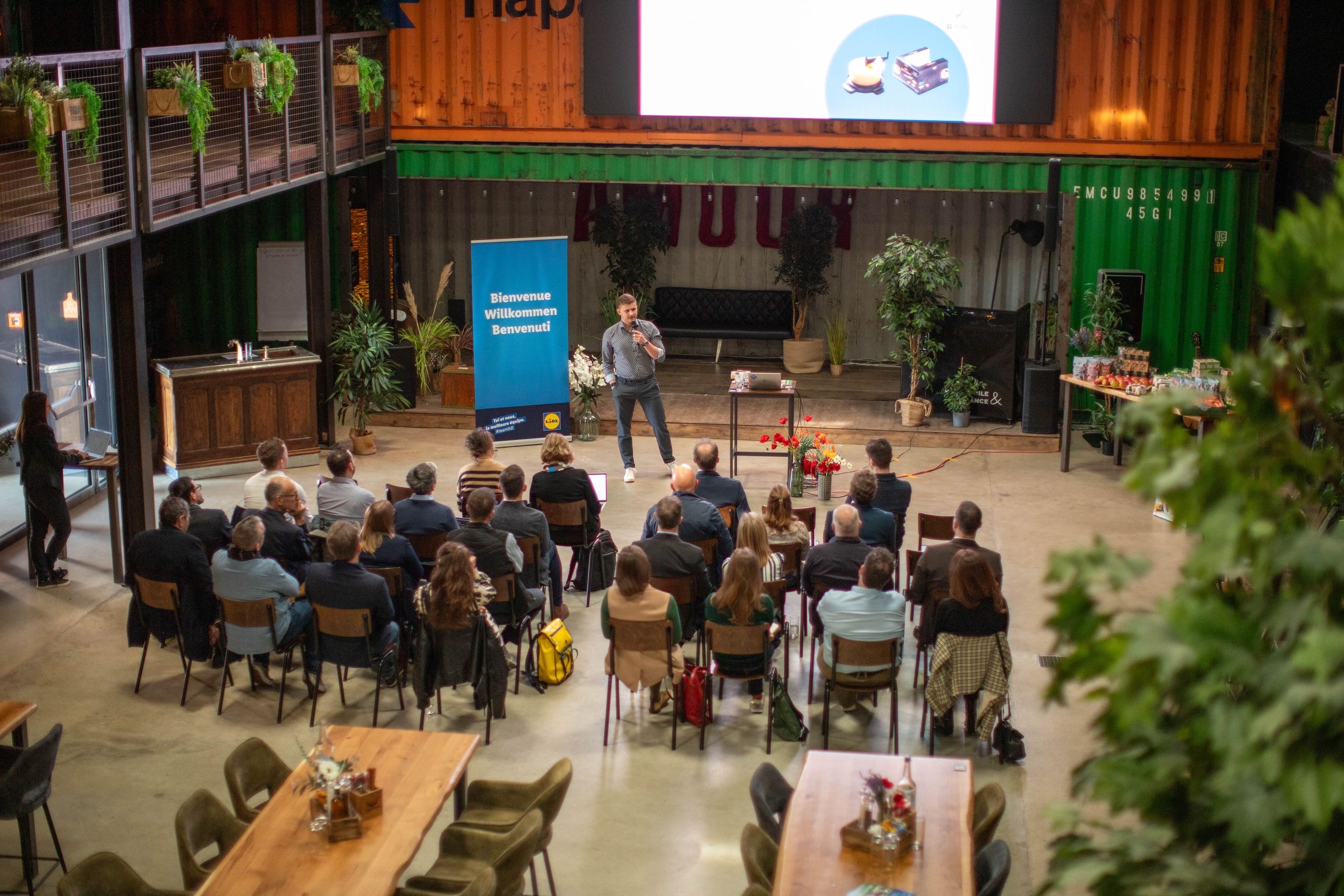 Ein Referent steht auf der Bühne und präsentiert vor dem Publikum, im Hintergrund der mehrsprachige Willkommensbanner von Lidl Schweiz.