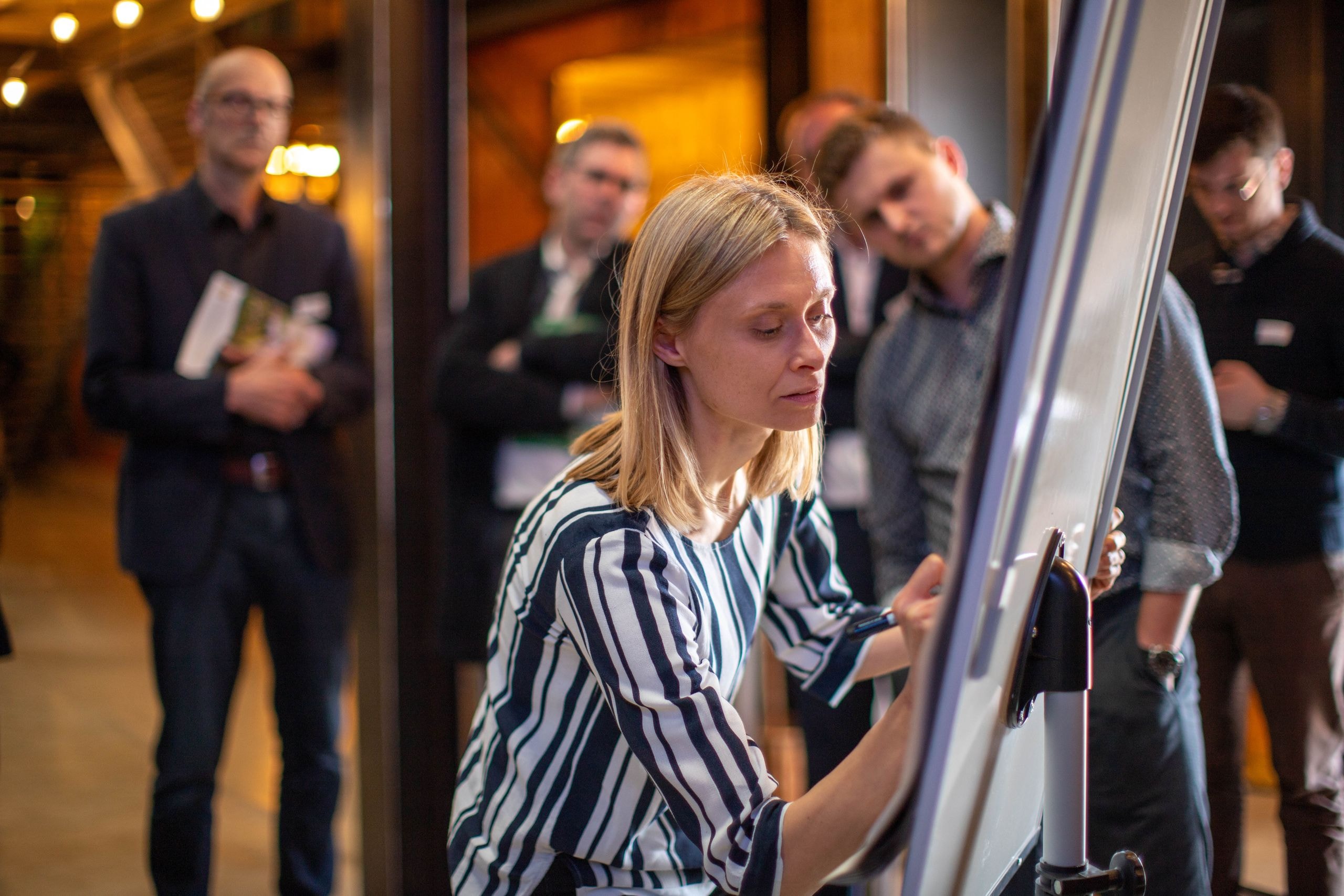 Eine Teilnehmerin gibt während einer interaktiven Session ihr Feedback auf einem Flipchart, während andere Gäste im Hintergrund diskutieren.