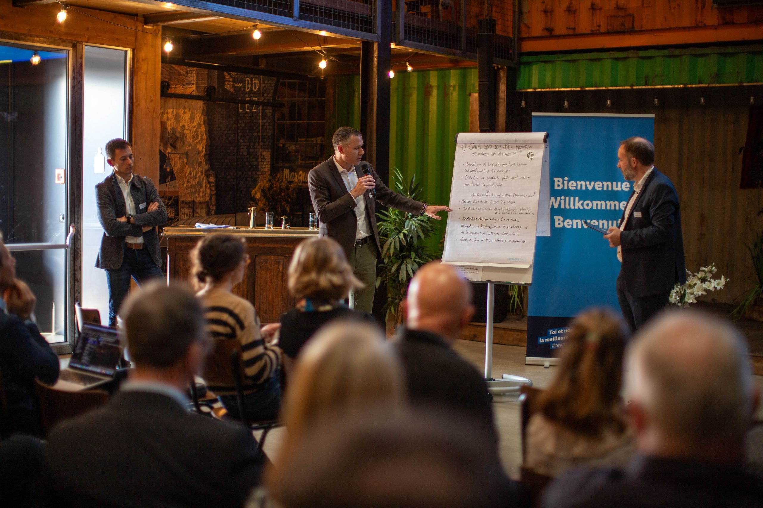 Drei Personen präsentieren die Ergebnisse einer Gruppenarbeit auf einem Flipchart vor dem Publikum des Stakeholder-Dialogs.