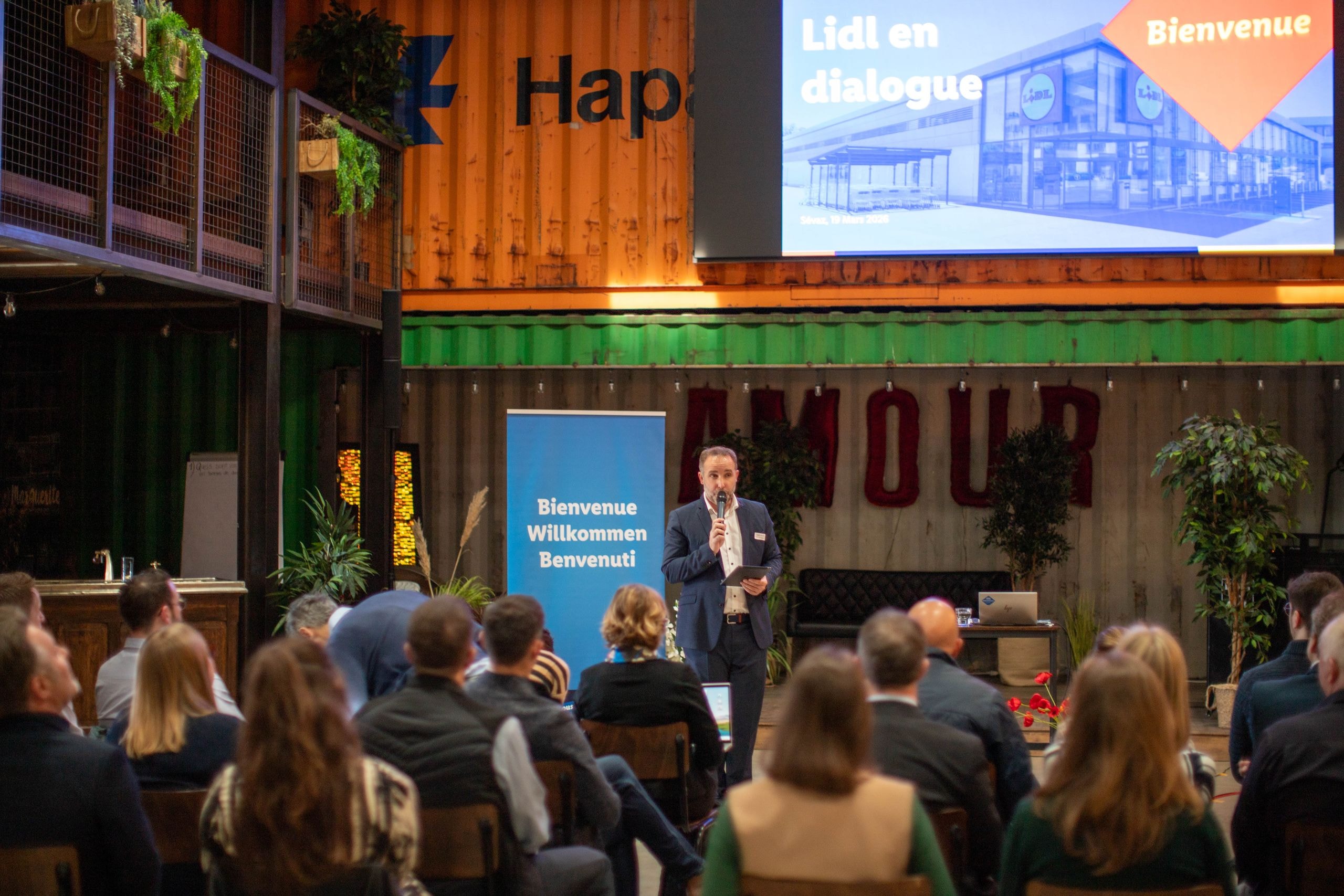 Ein Redner von Lidl Schweiz begrüsst die Gäste beim Stakeholder-Dialog, auf der Leinwand ist eine Lidl-Filiale mit dem Titel Lidl en dialogue zu sehen.