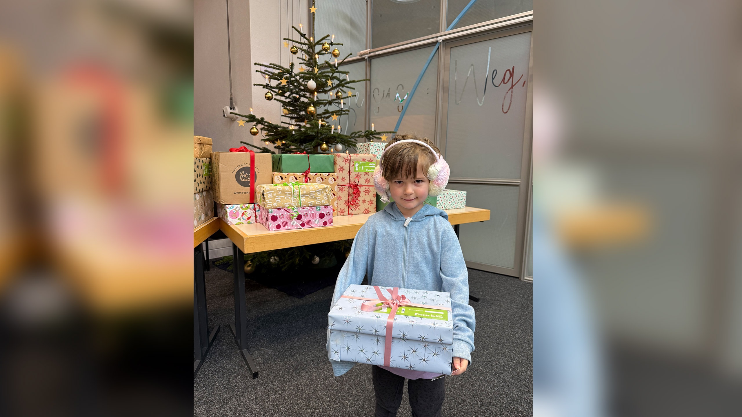 Ein Kind mit einem Weihnachtsgeschenk in den Händen steht vor einem Weihnachtsbaum und weiteren Geschenken.