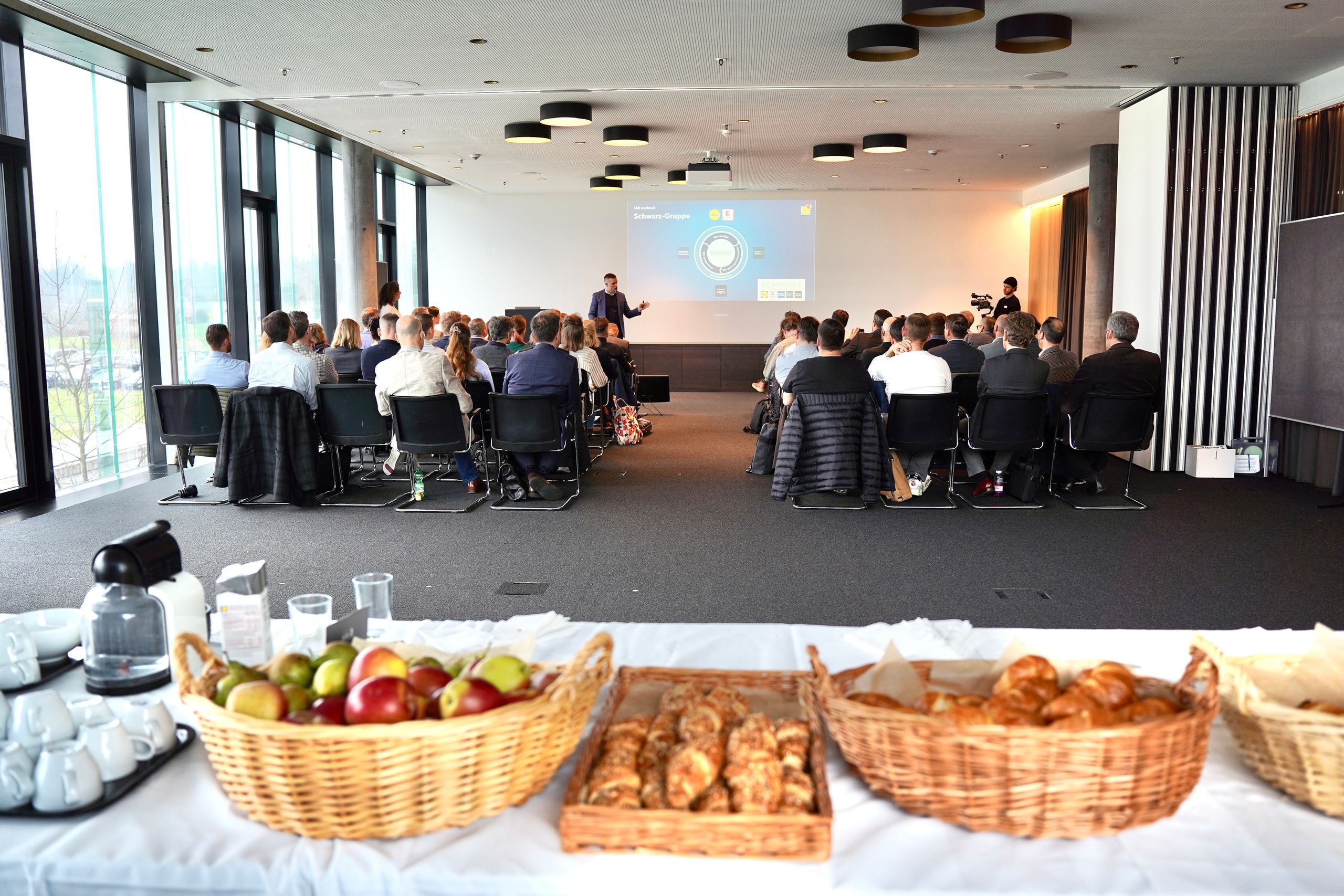 Vue d'ensemble de la réunion des parties prenantes de Lidl Suisse avec présentation du groupe Schwarz et table de restauration au premier plan.