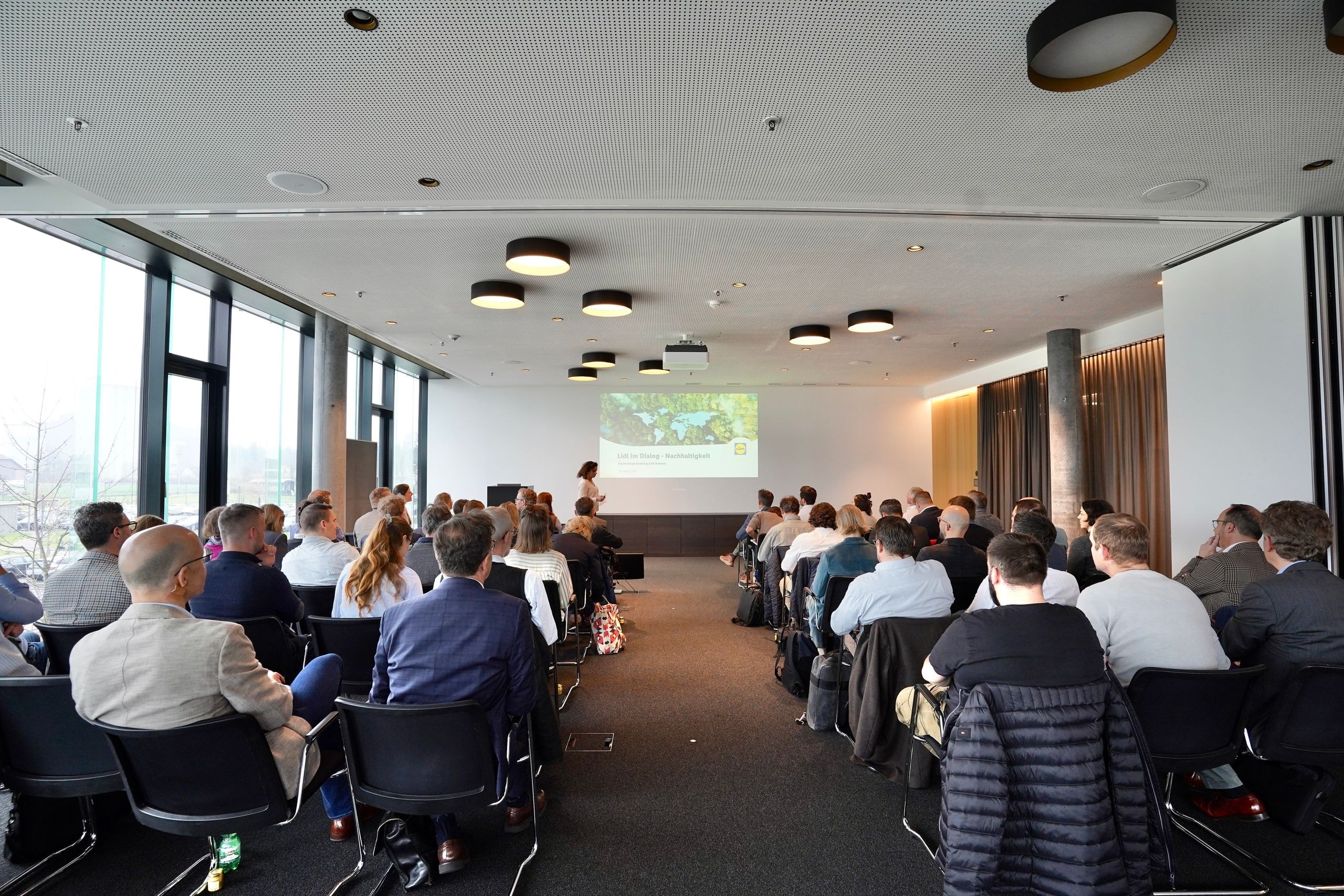 Dialogue avec les parties prenantes de Lidl Suisse sur le développement durable : une soixantaine de participants suivent une présentation sur le thème de la durabilité dans une salle de conférence moderne.